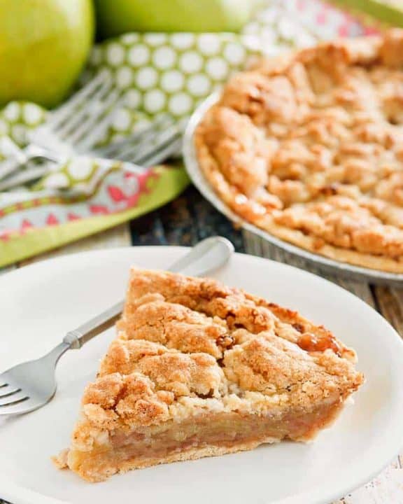 a slice of Dutch apple pie and a fork on a plate with the pie and apples in the background