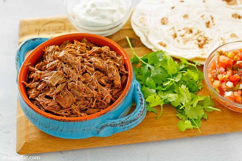 a bowl of shredded beef barbacoa, cilantro, sour cream, salsa, and tortillas