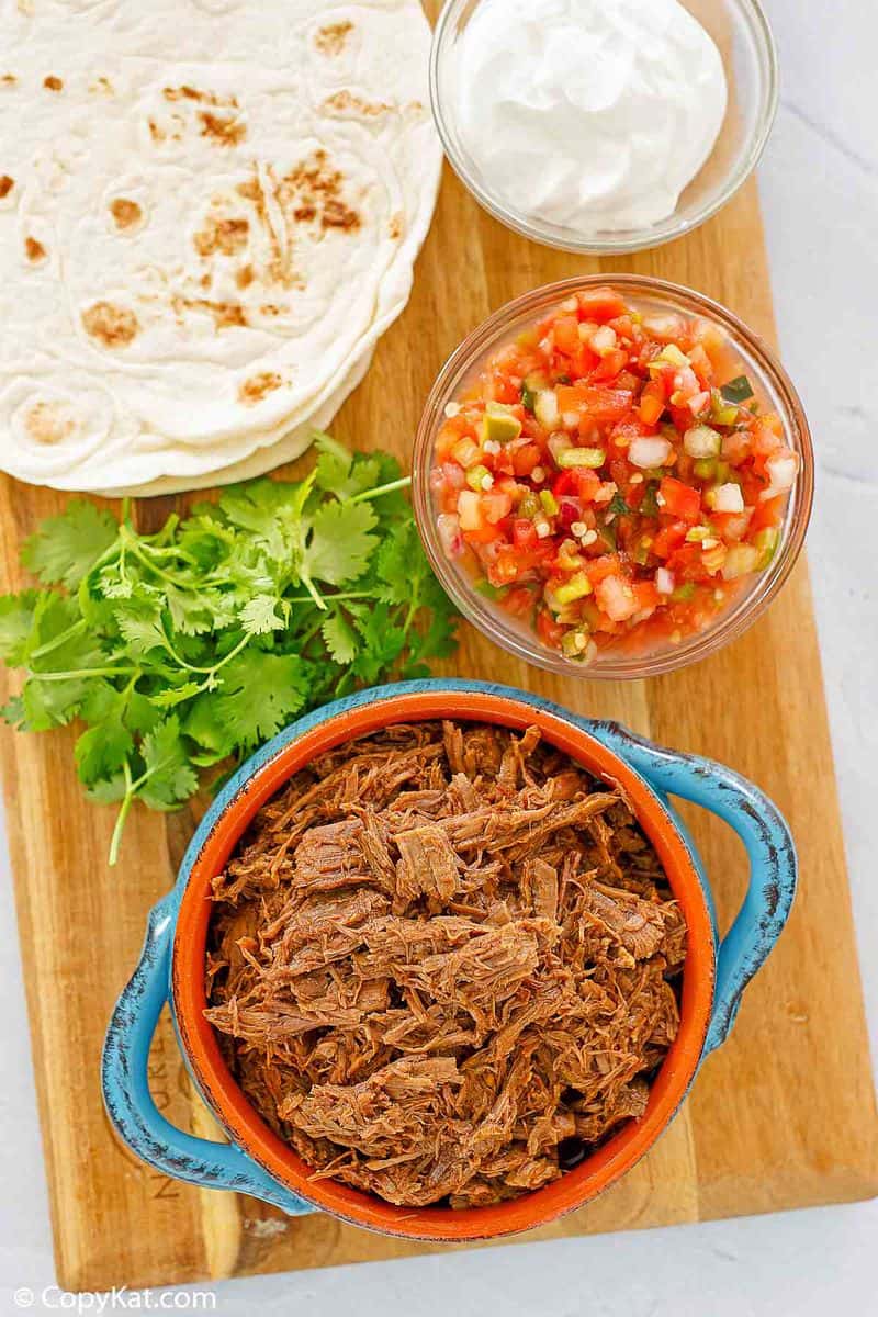 overhead view of beef barbacoa, cilantro, salsa, tortillas, and sour cream