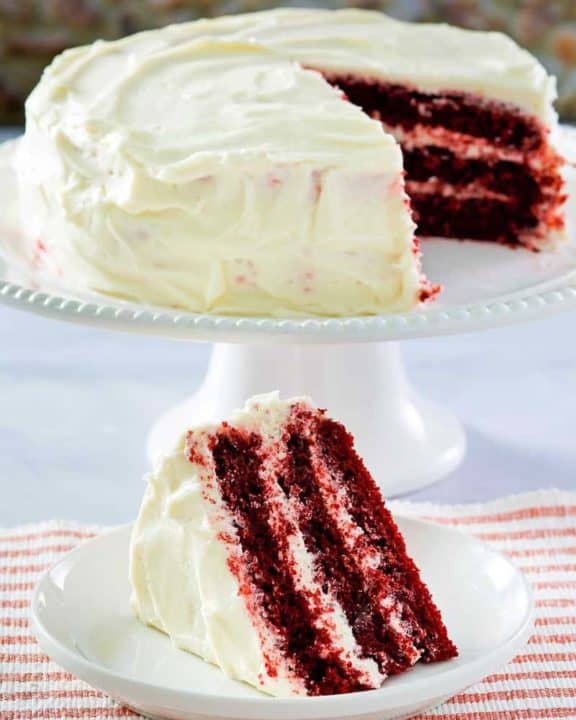 homemade Waldorf Astoria Red Velvet Cake slice on a plate in front of the cake on a stand
