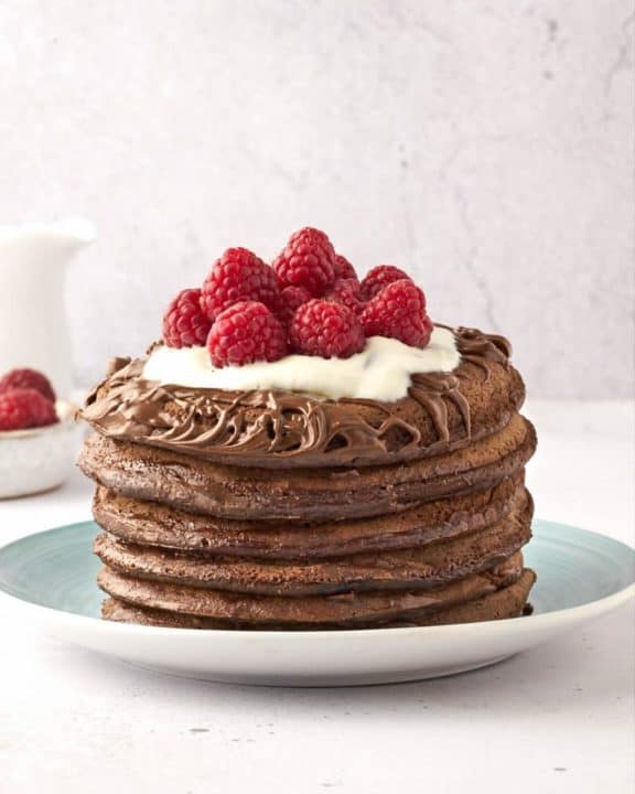 a stack of six chocolate pancakes topped with chocolate spread, whipped cream, and raspberries