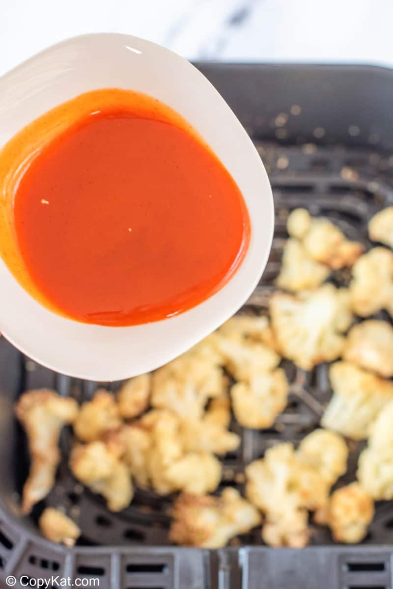 pouring buffalo sauce over cooked cauliflower in an air fryer basket