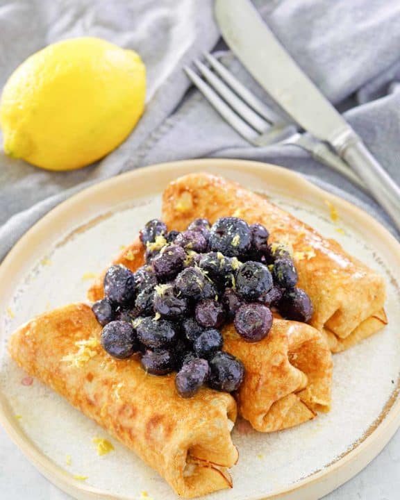 three cheese blintz topped with blueberries on a plate, a lemon, knife, and fork.
