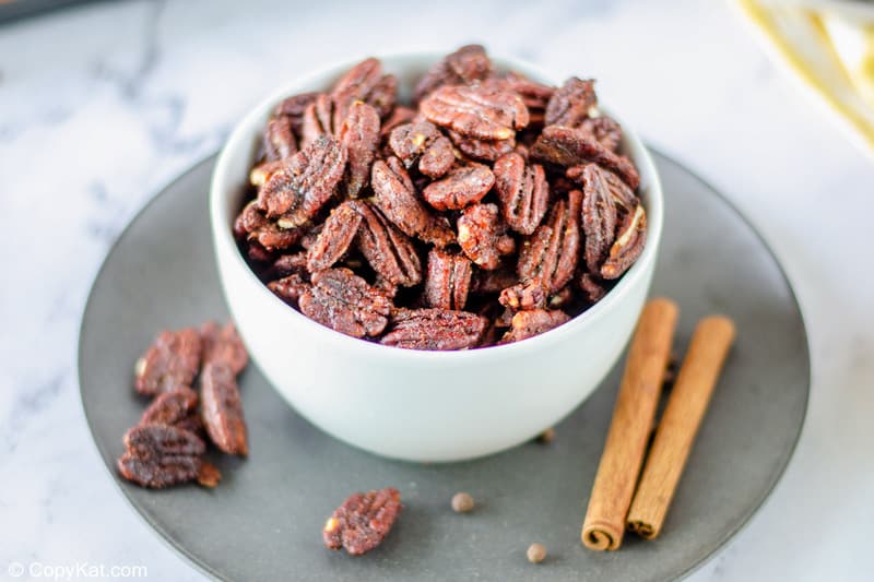 a bowl of spiced pecans and two cinnamon sticks on a plate