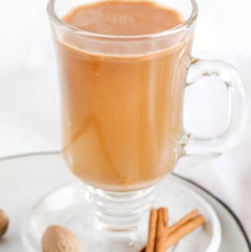 hot buttered rum in a glass mug on a plate with cinnamon sticks and nutmeg.