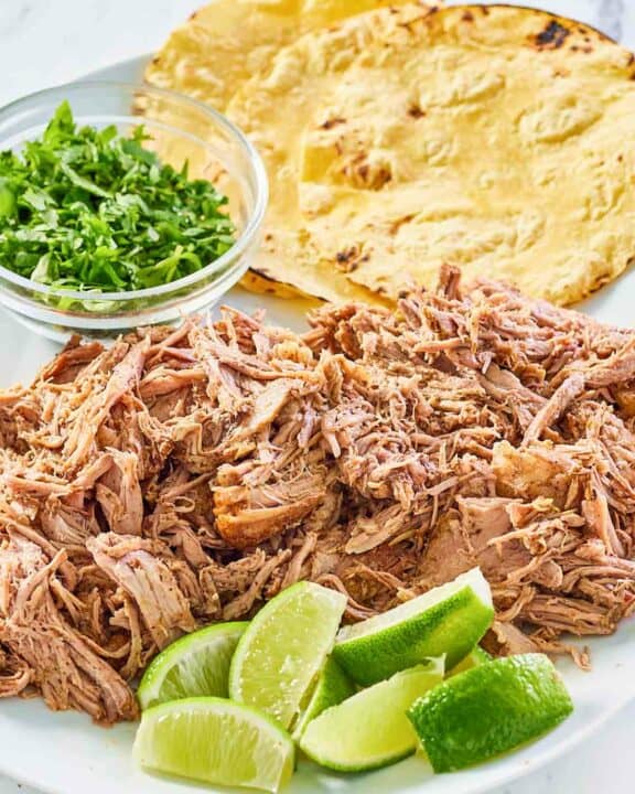pulled oven roasted pork shoulder on a platter with limes, cilantro, and tortillas.
