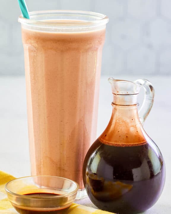 Homemade chocolate syrup in a small glass pitcher and a glass of chocolate milk.