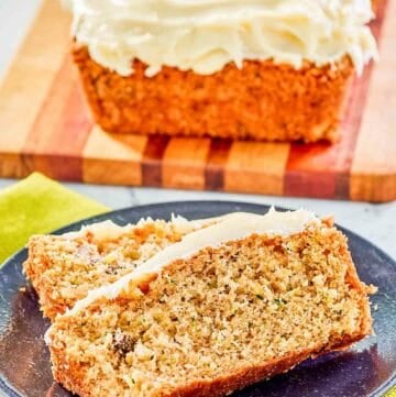 Two slices of zucchini nut bread with cream cheese frosting on a plate with a loaf behind it.