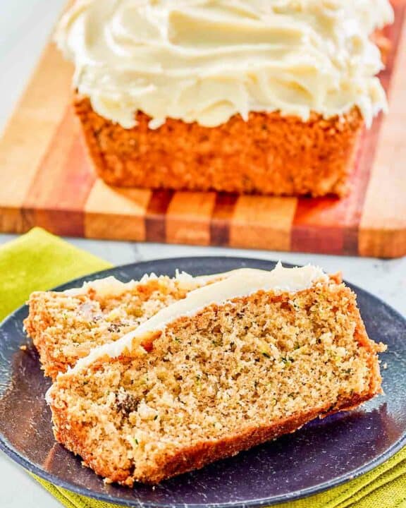 Two slices of zucchini nut bread with cream cheese frosting on a plate with a loaf behind it.