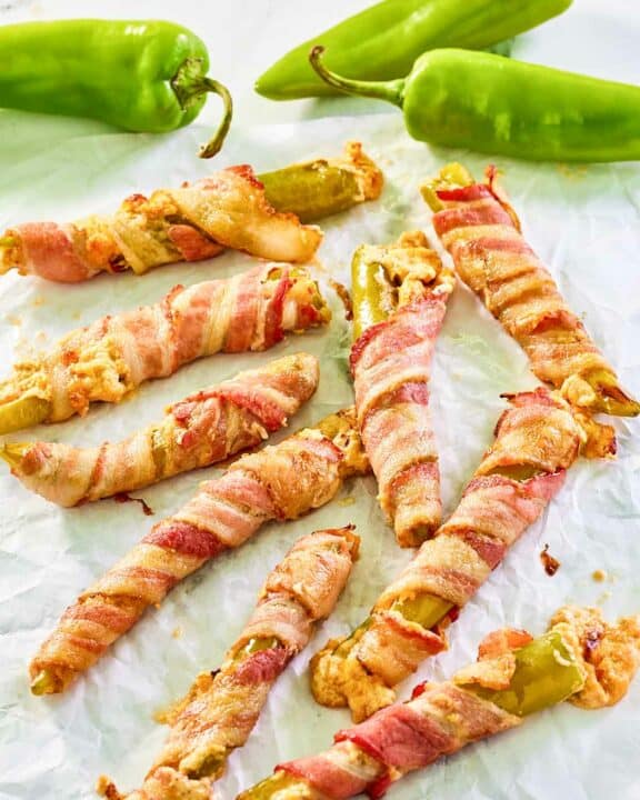 Bacon wrapped stuffed hatch peppers scattered on parchment paper.