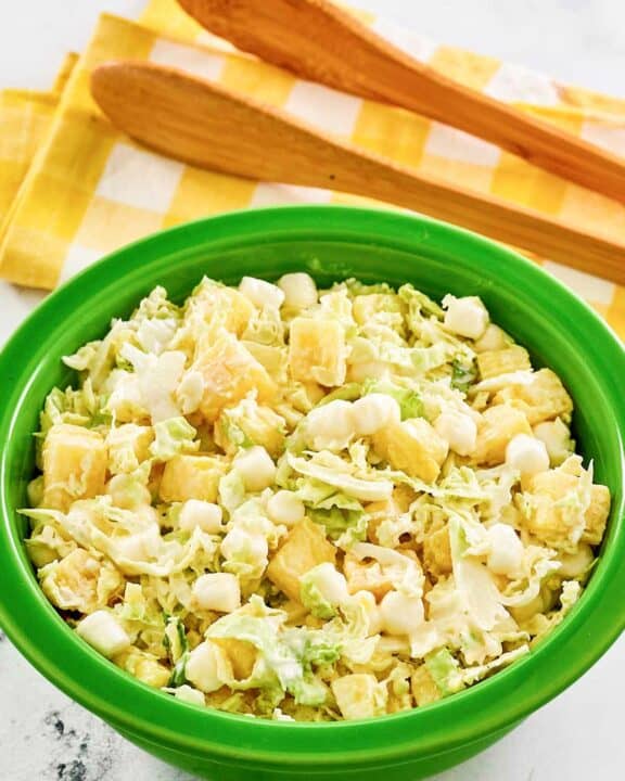 A bowl of pineapple coleslaw and salad tongs behind it.