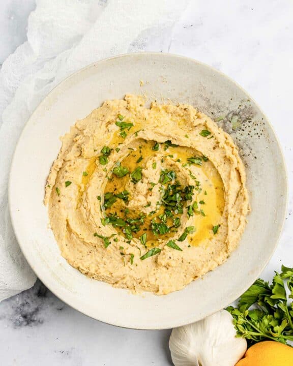 Overhead view of homemade garlic hummus, garlic bulb, lemon, and parsley.