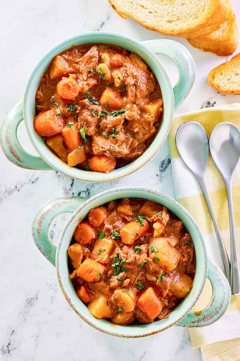 Crockpot beef stew with potatoes and carrots in bowls and spoons next to them.