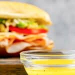 Homemade Mr. Submarine sub sauce in a small glass bowl and a sub sandwich behind it.