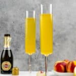 Two homemade Olive Garden peach bellini drinks, peaches, and bottle of sparkling wine.