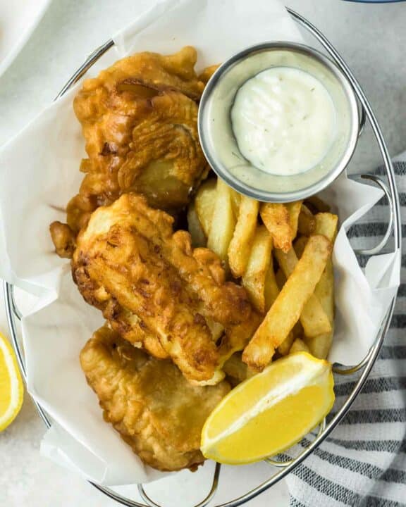 Beer battered fish and chips, lemon wedge, and a cup of tartar sauce in a serving basket.