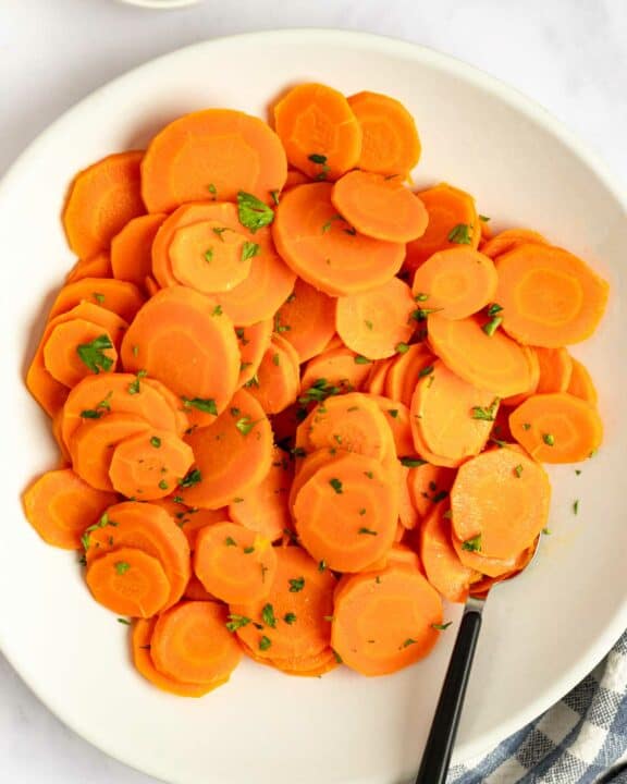 Cooked carrots on a plate and a bowl of chopped fresh parsley.