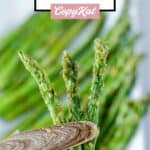 Wood tongs holding air fried asparagus over a platter of it.
