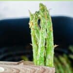 Wood tongs holding air fried asparagus over an air fryer.