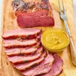 Baked corned beef, small bowl of mustard, and carving fork on a wood carving board.