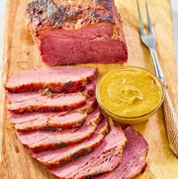 Baked corned beef, small bowl of mustard, and carving fork on a wood carving board.