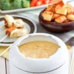 Copycat melting pot traditional swiss fondue in a fondue pot and dippers behind it.