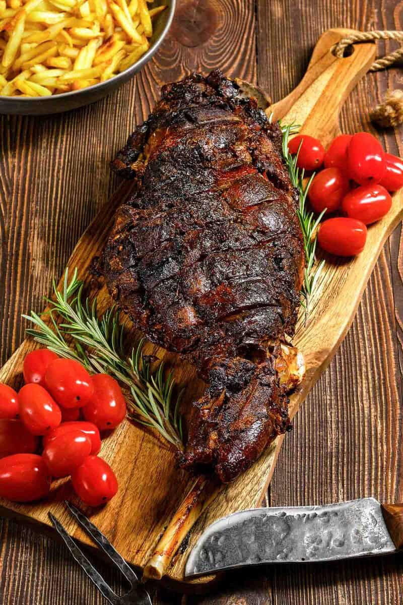 Oven roasted Indian style kashmiri leg of lamb, tomatoes, and rosemary on a wood board.