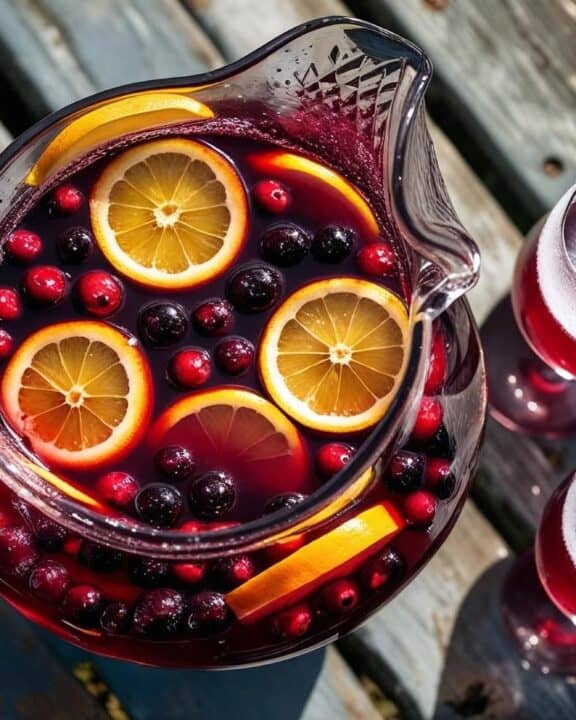Overhead shot of a glass pitcher filled with ruby‑red sangria dotted with orange wheels, lemon slices, and floating berries, set on a weathered picnic table beside frosty stemless wine glasses.