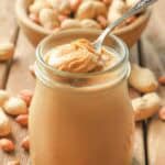 Homemade peanut butter in a glass jar and a bowl of peanuts behind it.